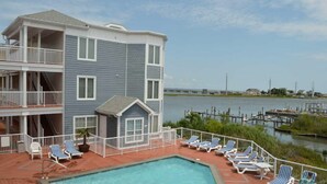 A multi-story building with a pool area featuring lounge chairs and a view of a body of water.