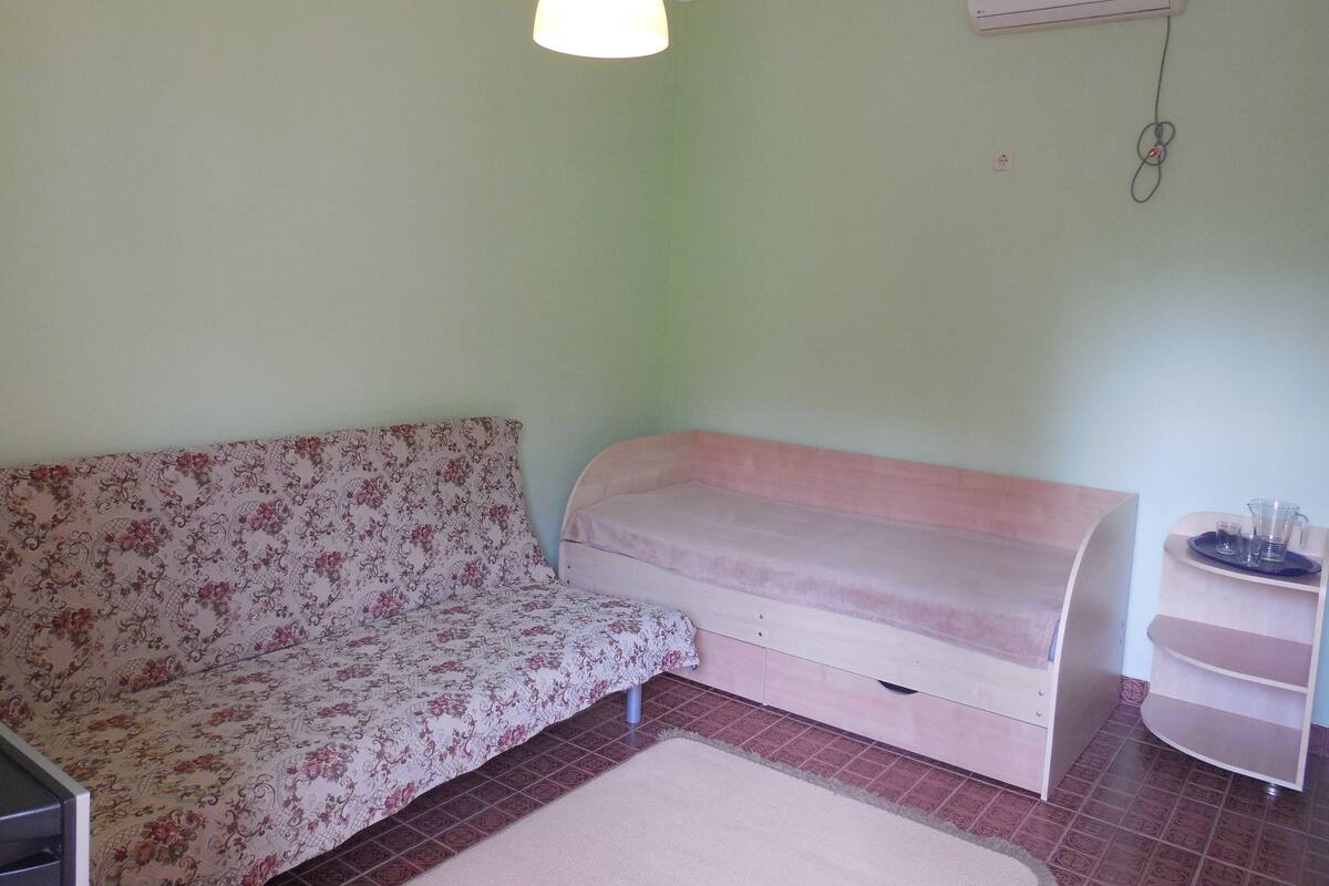 A small room with a floral-patterned sofa, a single bed, a small table with a glass, and a wall-mounted air conditioner.
