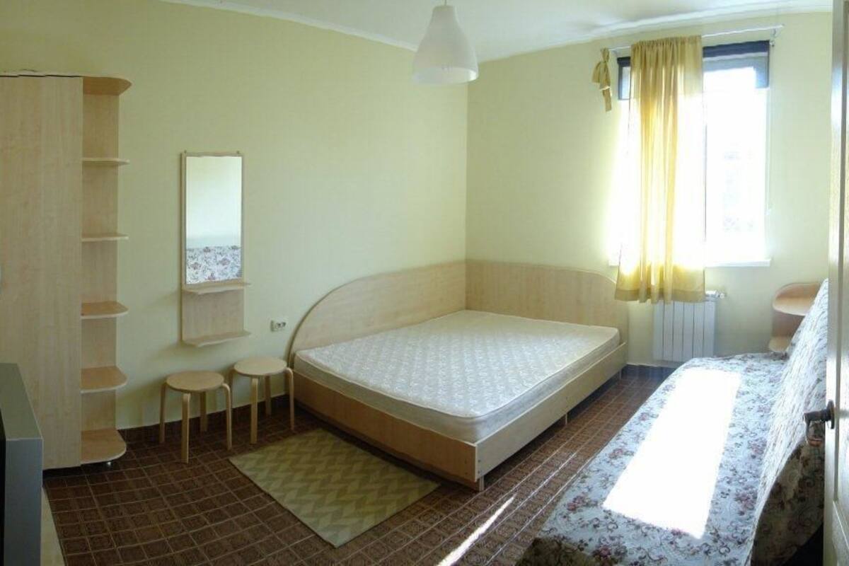 A bedroom with a single bed, a wardrobe, a small table, a chair, and a window with curtains.