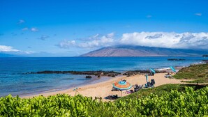 A beach with clear blue water, a sandy shore, and a mountain in the background. There are umbrellas and chairs on the beach. - Kamaole Sands 3-406 - Modern Kitchen, Large Lanai, Enclosed Private MB, Split AC (Kihei)