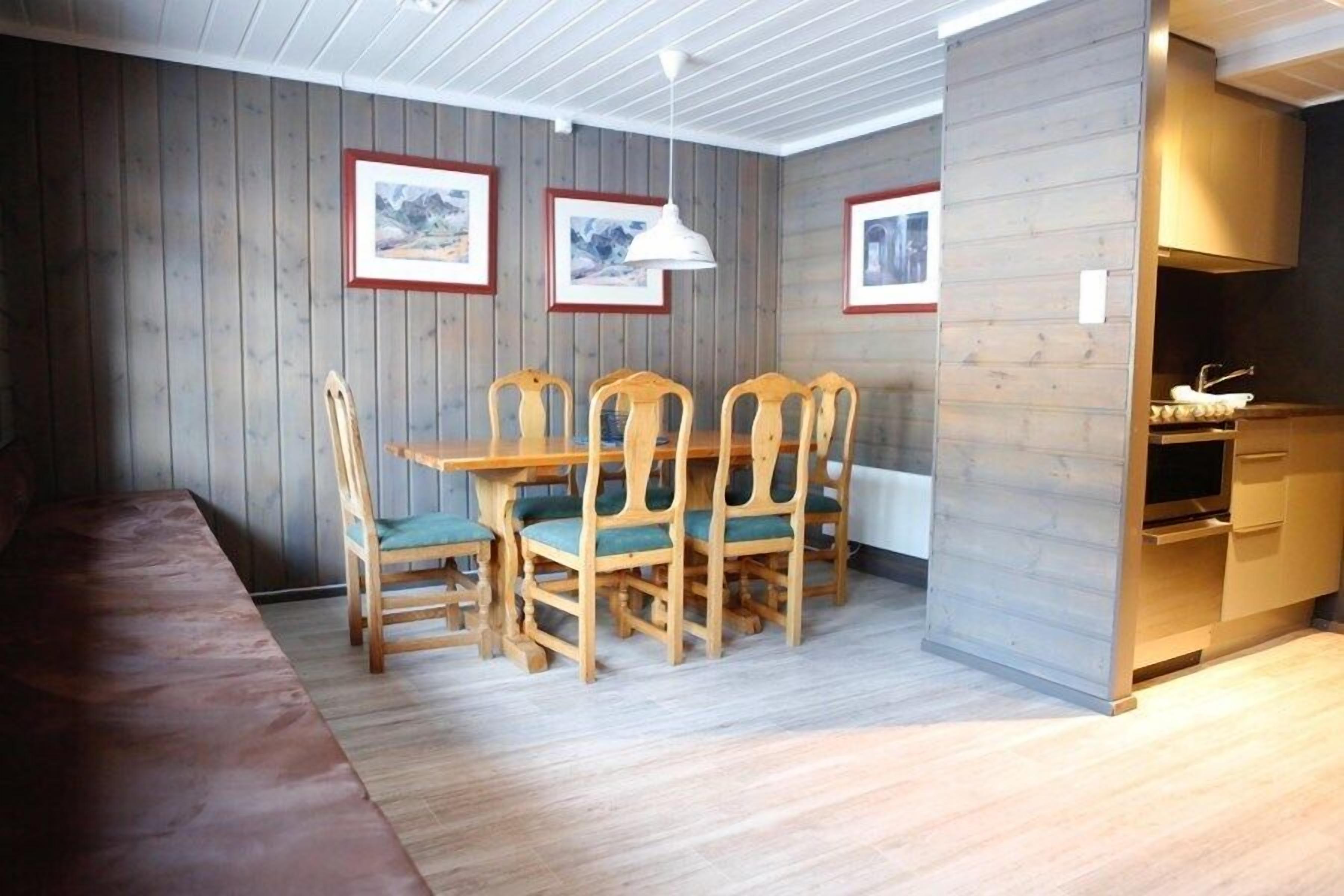 A dining area with wooden chairs and a table, framed pictures on the wall, and a kitchen area with wooden cabinets.