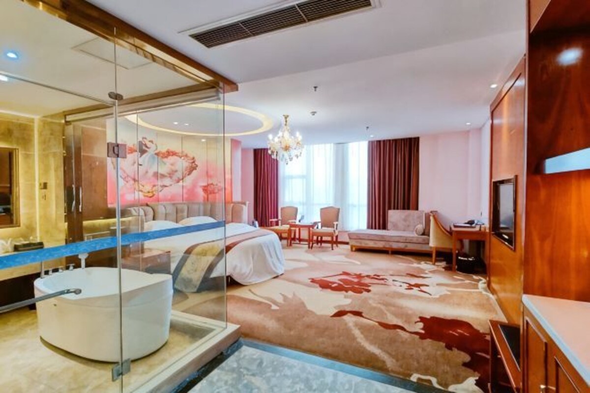 A hotel room with a glass-enclosed bathroom, a large bed, a seating area with a sofa and chairs, and a television.