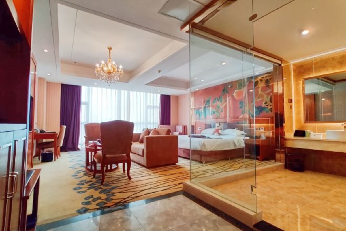 A hotel room with a bed, a seating area with chairs, a dining table, a chandelier, and a bathroom with a mirror and sink.
