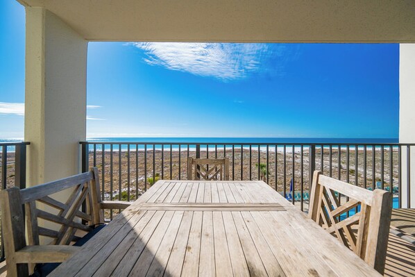 A wooden deck with a table and chairs, overlooking a beach with a clear sky.