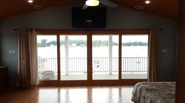 A spacious room with a large sliding glass door leading to a balcony, wooden flooring, a bed with a patterned bedspread, and a ceiling fan.