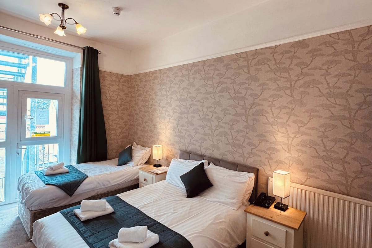 A hotel room with two beds, a window with curtains, a radiator, and a nightstand with a lamp.