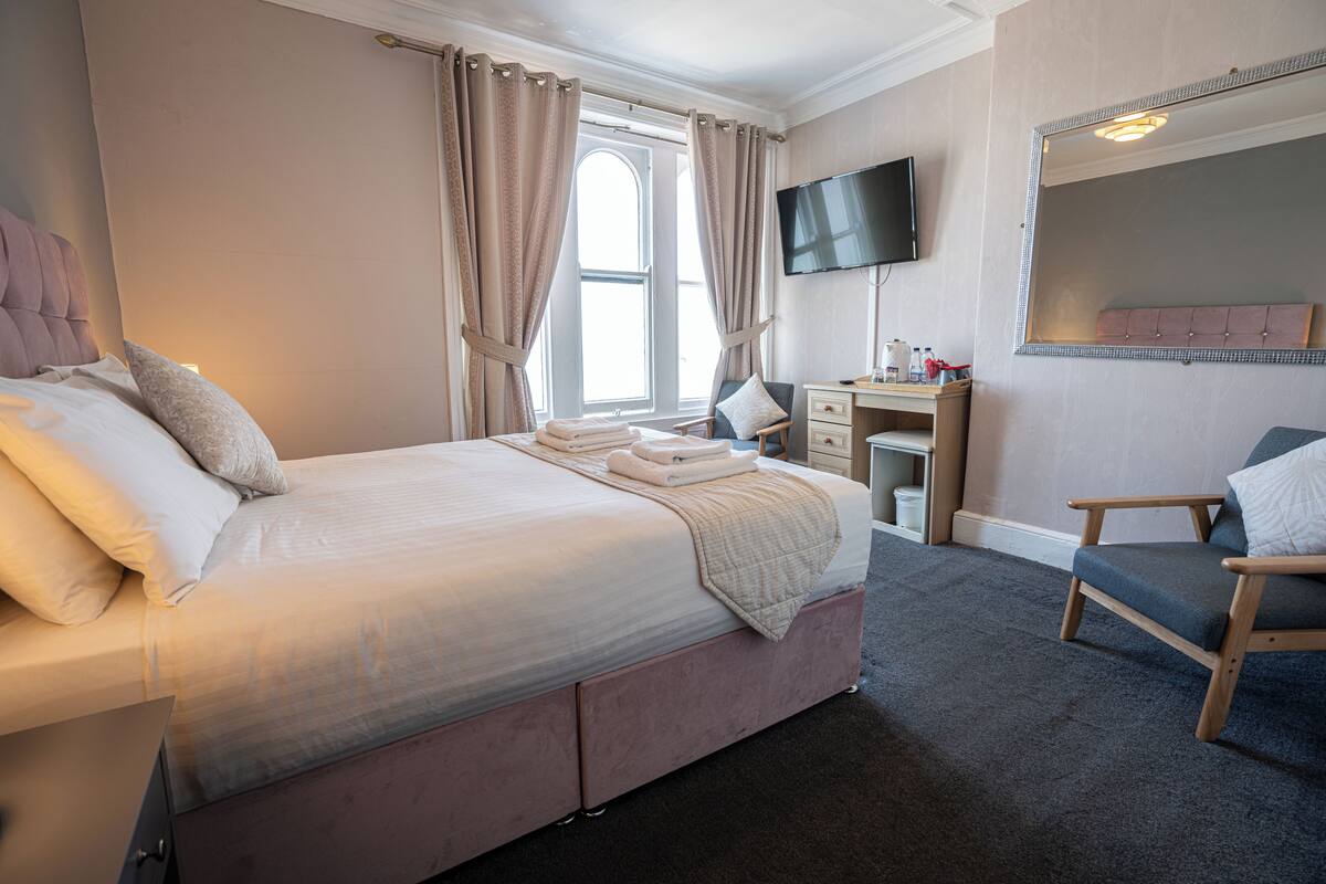 A hotel room with a large bed, a desk, a chair, a TV, and a window with curtains.