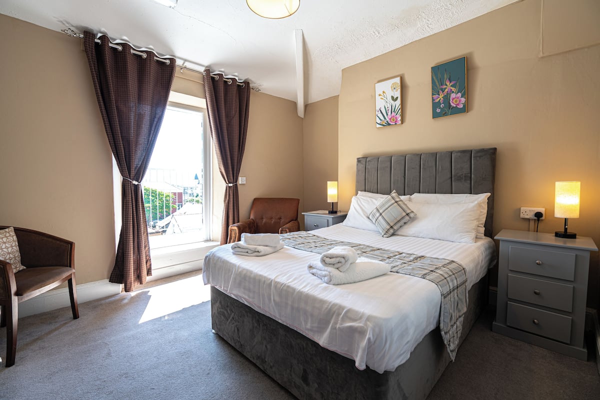 A hotel room with a large bed, a chair, a nightstand, and a window with curtains.