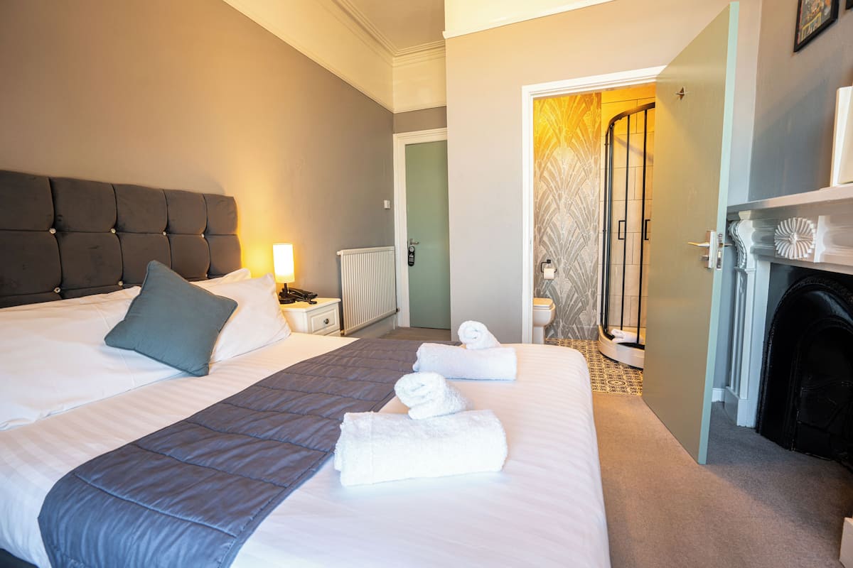 A neatly made bed with a grey quilt, white linens, and two rolled towels. There is a bedside table with a lamp and a radiator in the background.