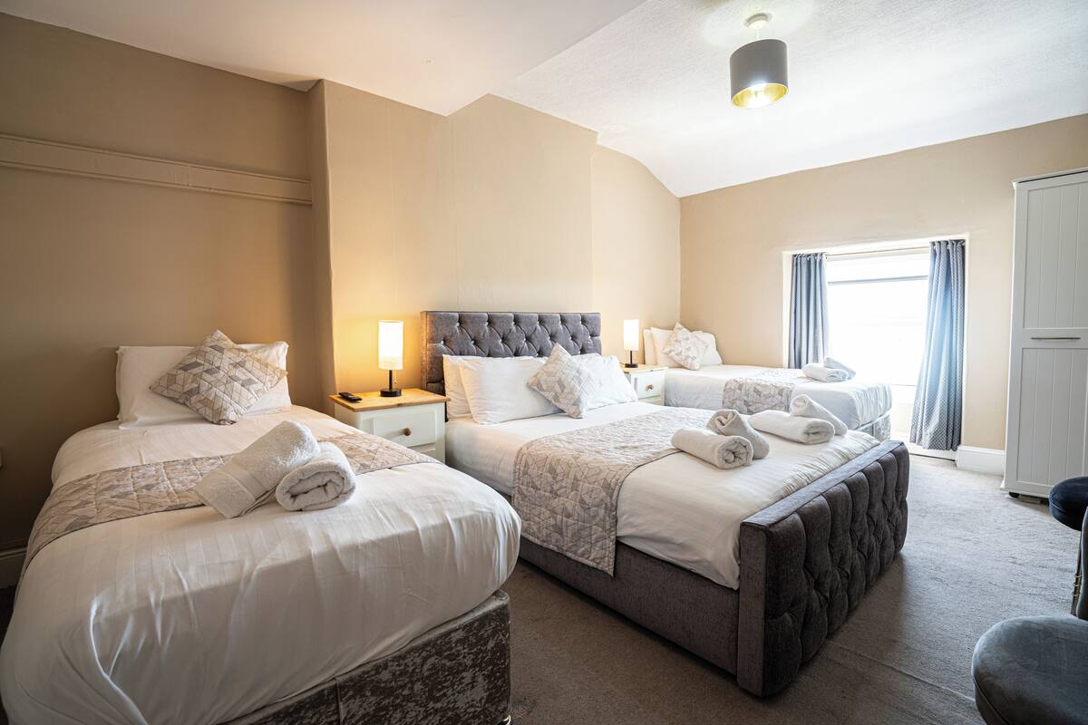 A hotel room with two beds, a large window with curtains, a wardrobe, and a bedside table with a lamp.