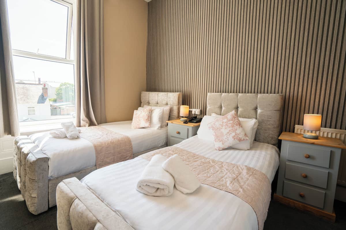 A hotel room with two beds, a nightstand with a lamp, and a window with curtains.