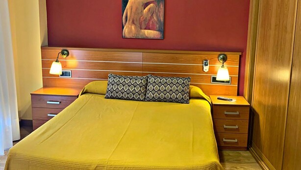 A hotel room with a large bed, wooden headboard, two bedside tables with lamps, a red accent wall, and a painting above the bed. - Hostal Doña Juana (Ávila)