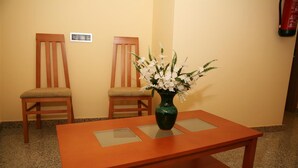 A wooden table with a vase of white flowers, two wooden chairs with cushions, and a wall-mounted electrical outlet. - Hostal Doña Juana (Ávila)