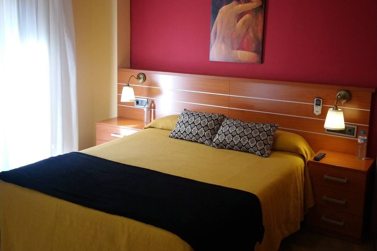 A hotel room with a bed, a nightstand, a lamp, a painting, and a window with curtains.