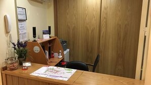 A reception desk with a wooden finish, a potted plant, a glass jar, and a printed document on the counter. - Hostal Doña Juana (Ávila)