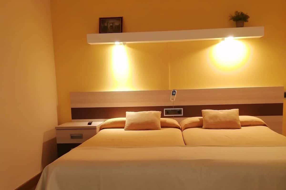 A hotel room with a bed, bedside tables, a nightstand, a wall-mounted shelf with a plant, and a wall-mounted light fixture.