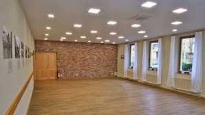Een ruime kamer met een houten vloer, bakstenen muur en grote ramen met witte gordijnen.