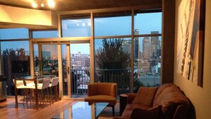 Ein modernes Wohnzimmer mit einem Glastisch, einem braunen Sofa und einem Essbereich mit weißem Tisch und Stühlen. Große Fenster bieten Blick auf die Stadtlandschaft.