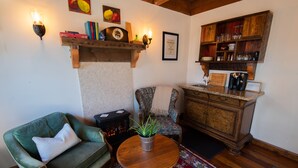 A cozy living room with a green armchair, a wooden coffee table with a plant, a fireplace, and a wooden cabinet with glassware.
