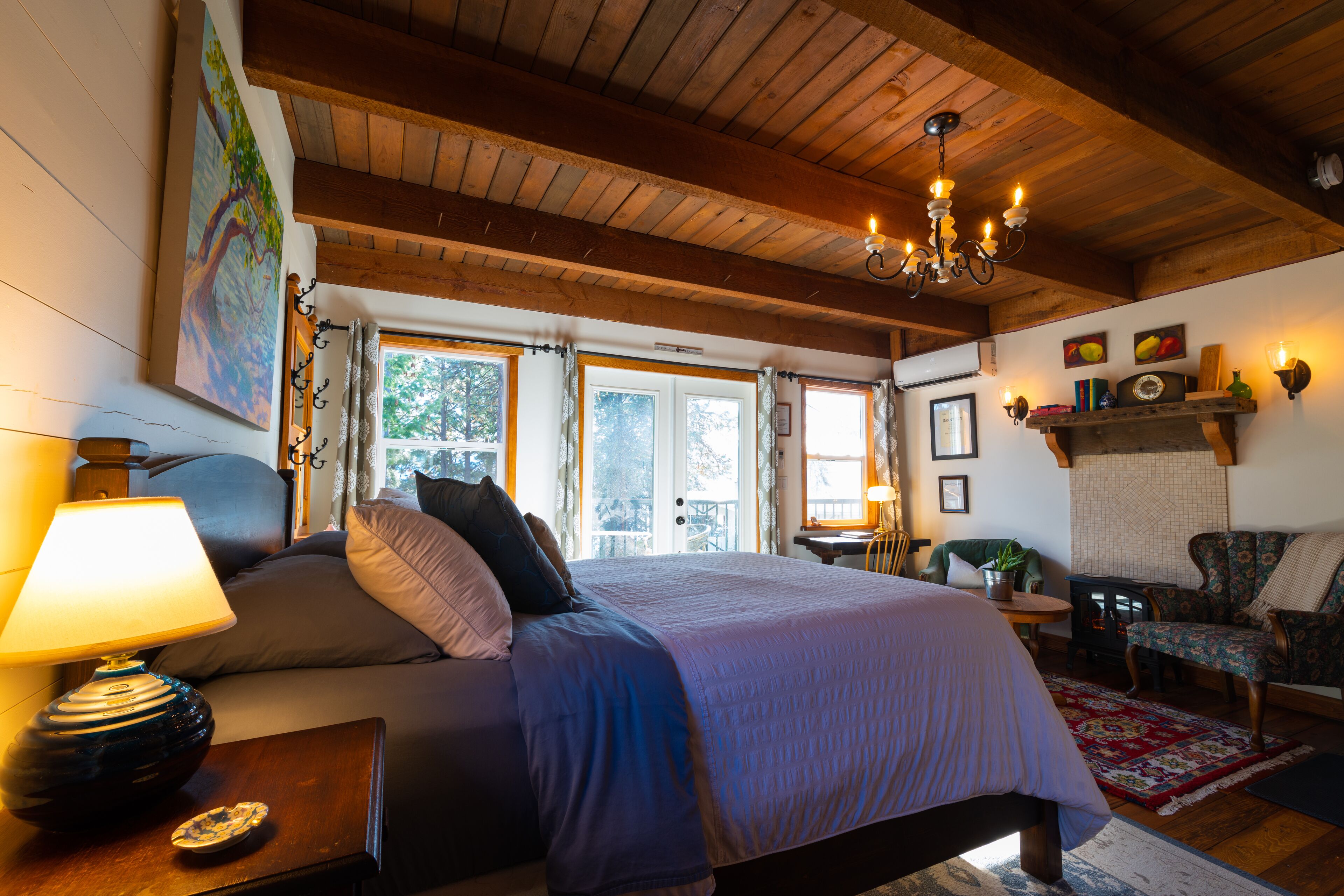 Une chambre avec un grand lit, un plafond en bois, un lustre, une fenêtre avec des rideaux, une chaise et une petite table avec une lampe.