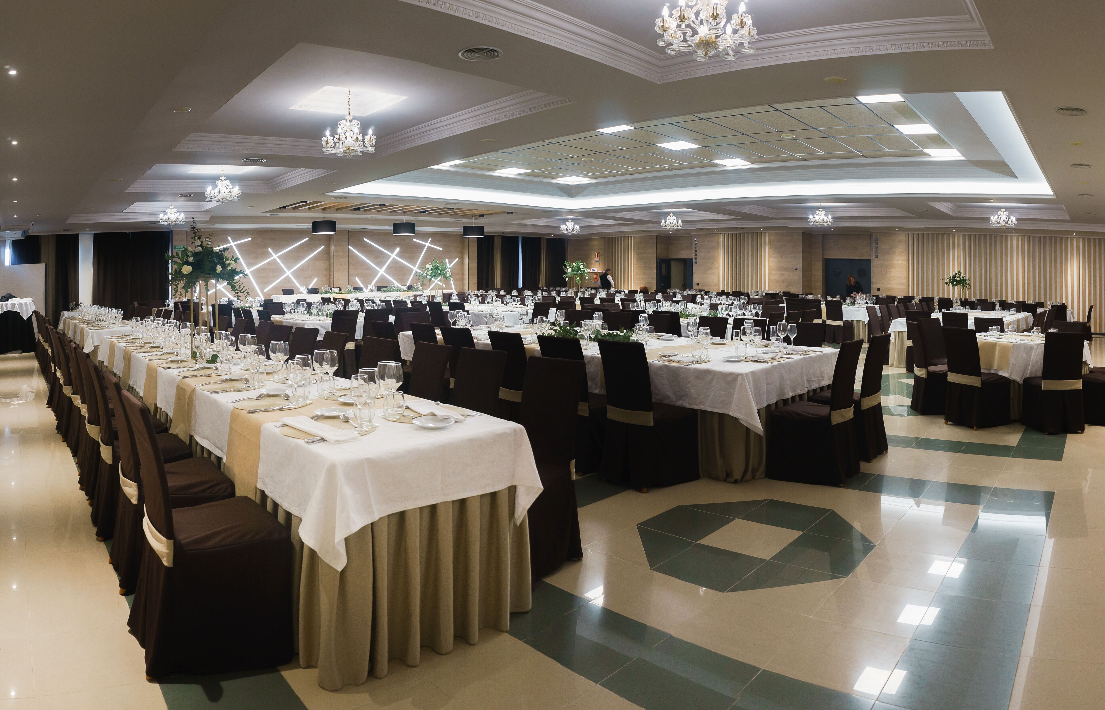 Un salón de banquetes con varias mesas redondas dispuestas para comer, con manteles blancos y sillas oscuras.