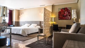 Habitación de hotel con cama, sofá, escritorio y silla. Hay un cuadro en la pared y una lámpara en el escritorio.