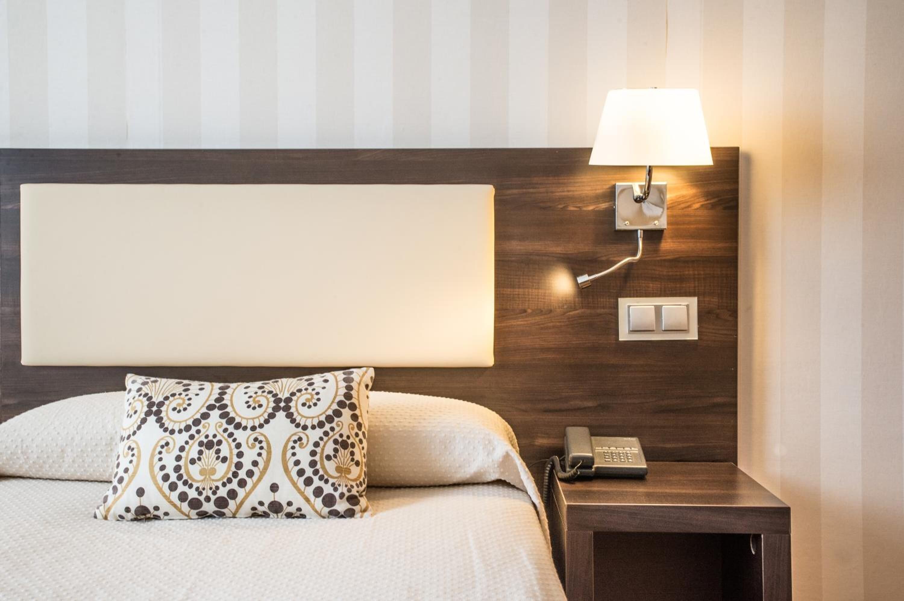 A neatly made bed with a patterned pillow, a bedside lamp, a phone, and a wall-mounted light switch.
