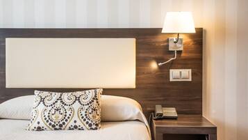 A neatly made bed with a patterned pillow, a bedside lamp, a phone, and a wall-mounted light switch.