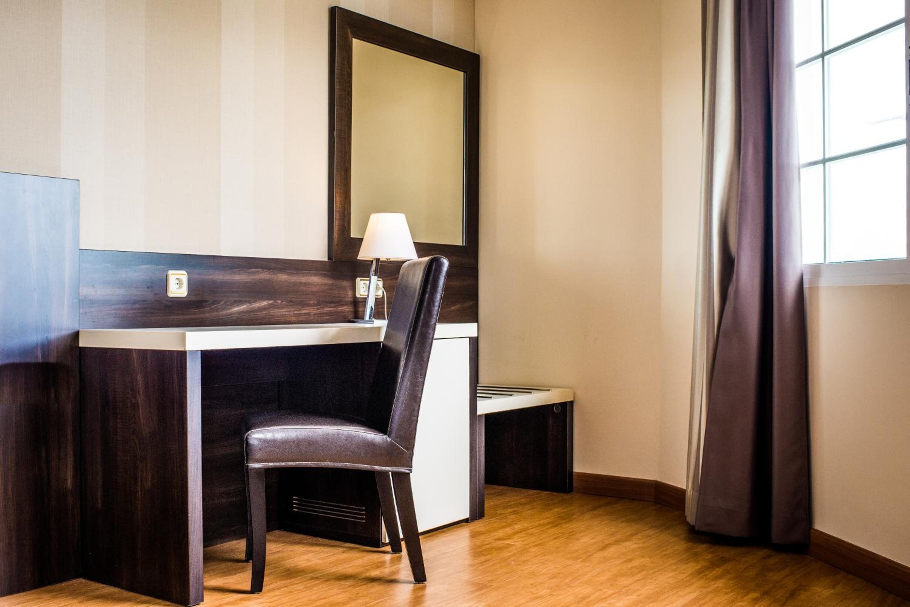 Habitación de hotel con piso de madera, un escritorio con silla, una lámpara y un espejo grande en la pared.