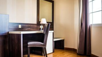 Habitación de hotel con piso de madera, un escritorio con silla, una lámpara y un espejo grande en la pared.