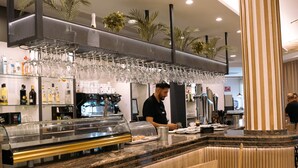 Zona de bufé con vitrina de vidrio, mostrador con cafetera y área de asientos con un pilar a rayas.