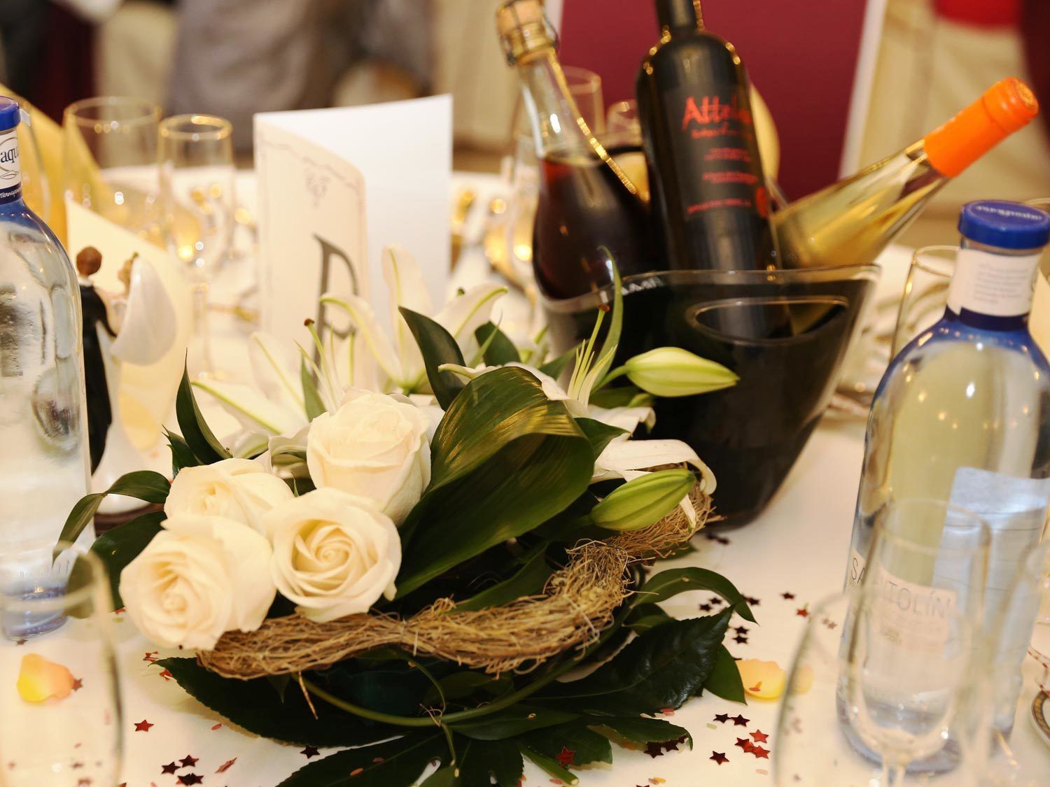 Una mesa con un ramo de rosas blancas, una cesta y varias botellas de vino.