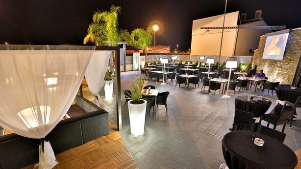 Un área de comedor al aire libre con mesas y sillas, un toldo blanco y plantas en macetas.
