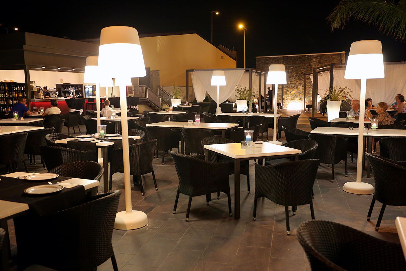 An outdoor dining area with tables and chairs, illuminated by large lamps.