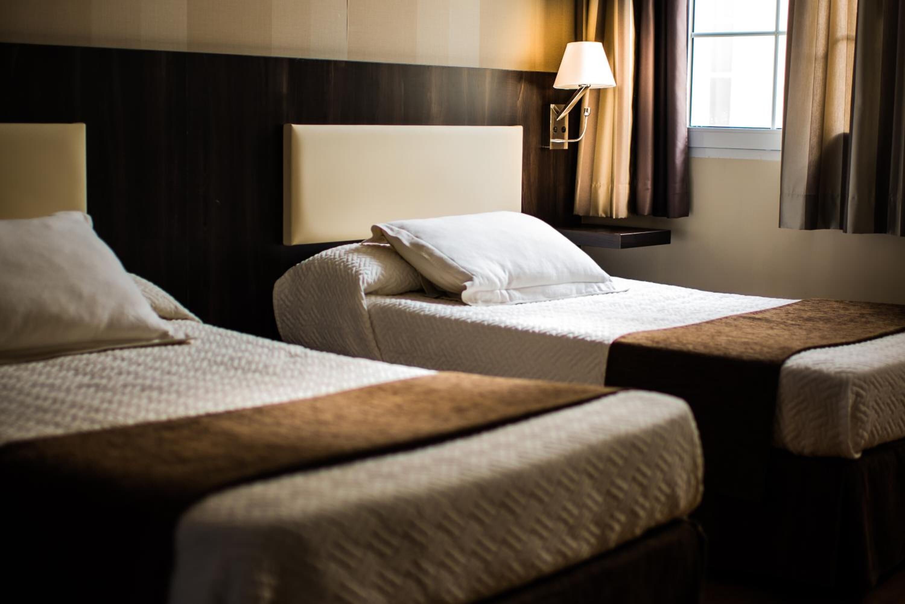 Una habitación de hotel con dos camas individuales, una mesita de noche, una lámpara y una ventana con cortinas.