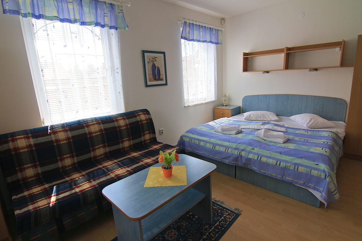 A bedroom with a bed, a sofa, a small table with a plant, and two windows with curtains.
