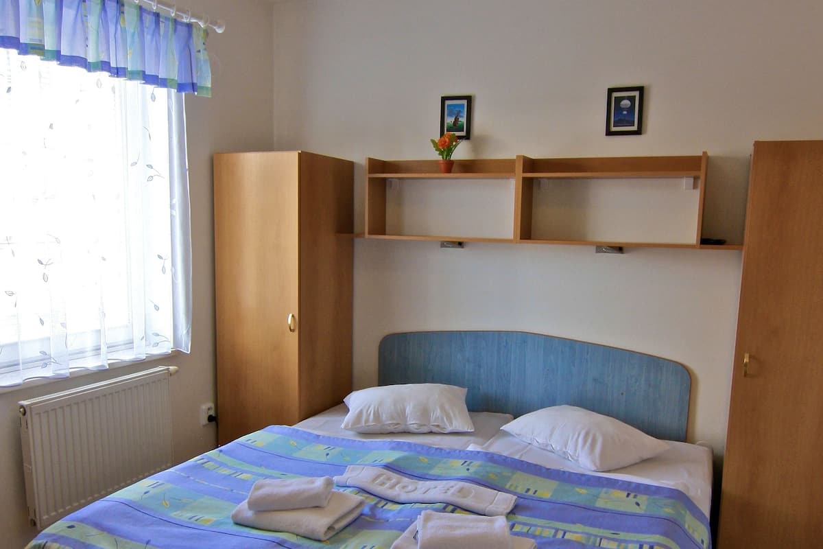 A bedroom with a bed, a window with curtains, a radiator, wooden wardrobe, and a shelf with pictures.