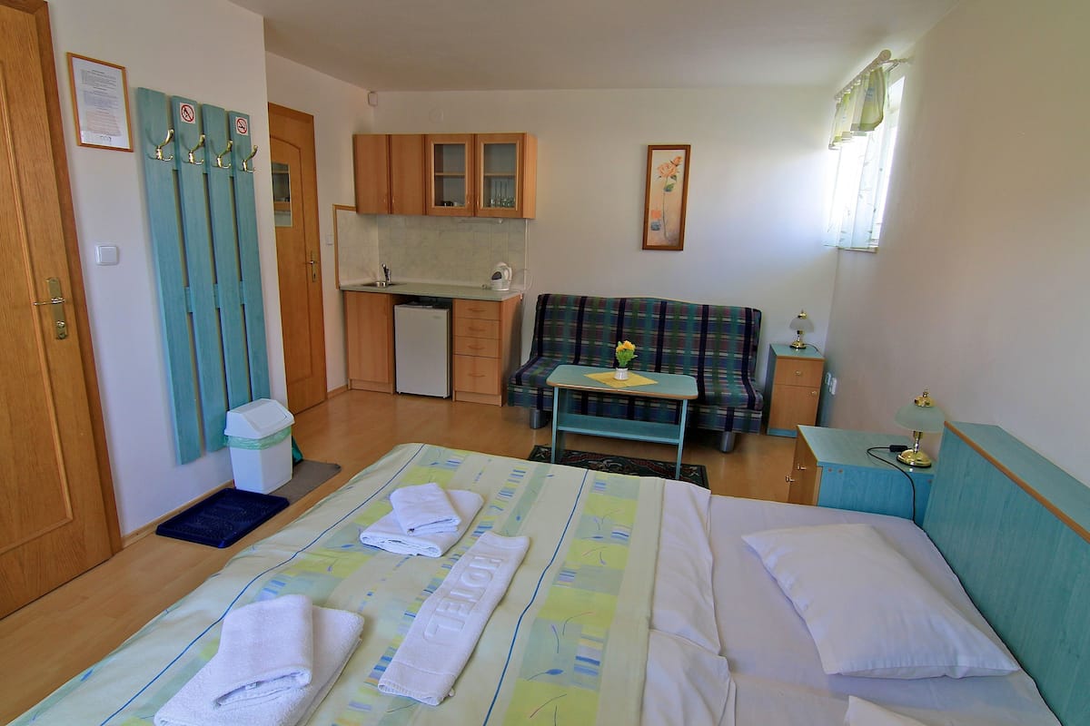 A hotel room with a bed, a sofa, a small table, a desk, a kitchen area, and a window with curtains.