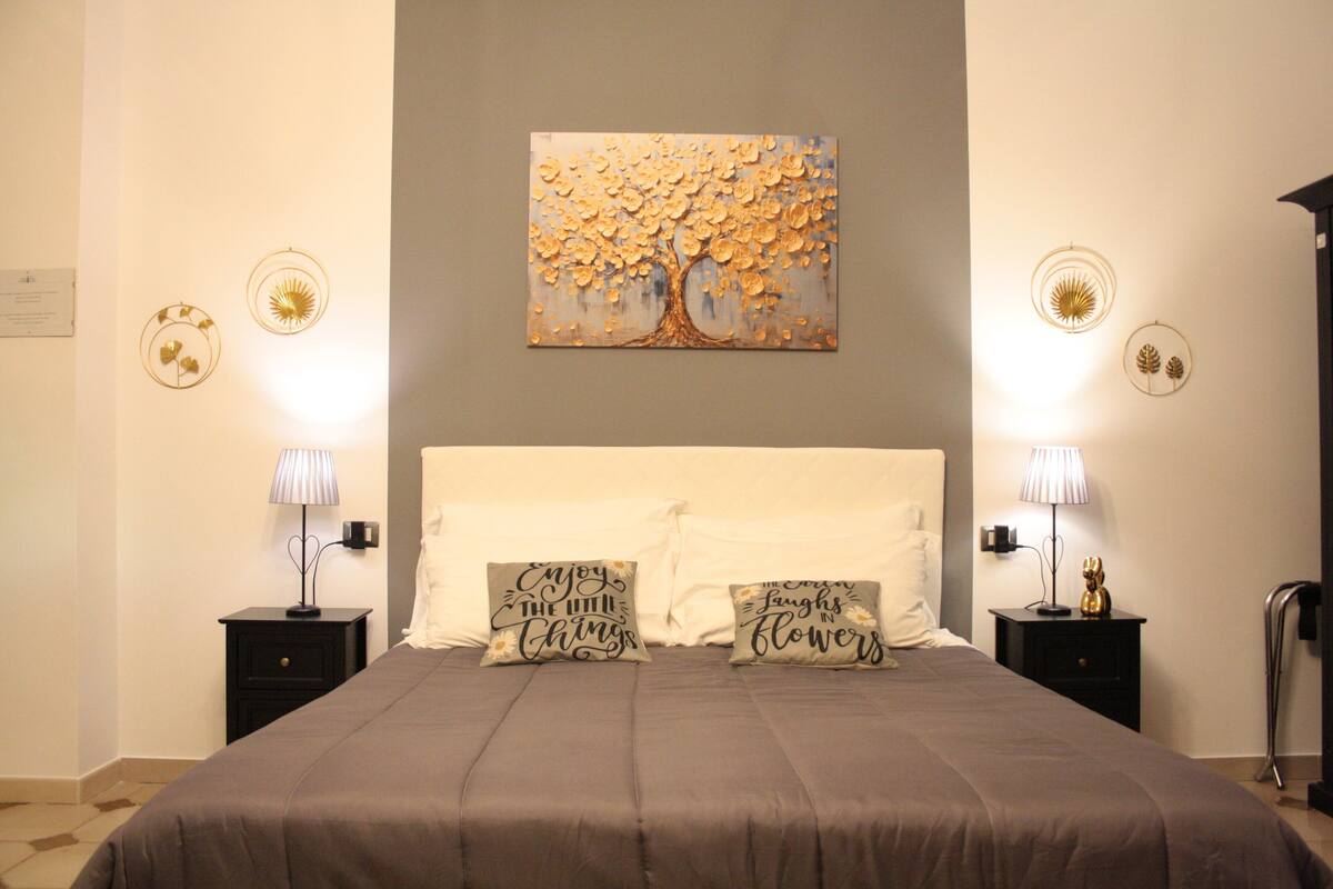 A bedroom with a large bed, two bedside tables with lamps, a wall-mounted painting of a tree, and decorative wall art.
