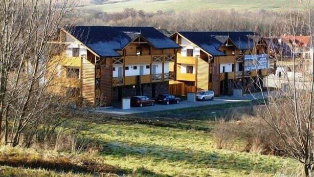 A row of wooden cabins with sloped roofs, arranged in a linear fashion with a grassy field and trees in the background.