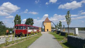 A red tram at a station with a yellow building and a bridge in the background.