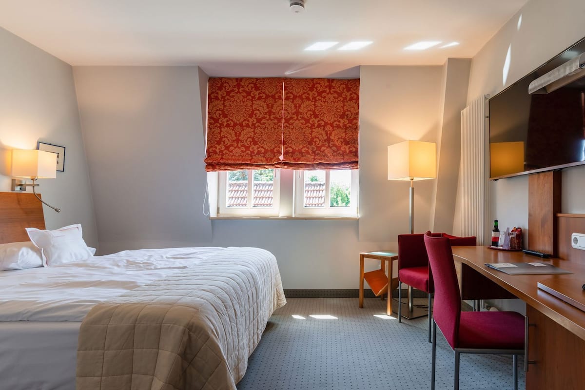 A hotel room with a bed, a desk, a chair, a TV, and a window with a red valance.