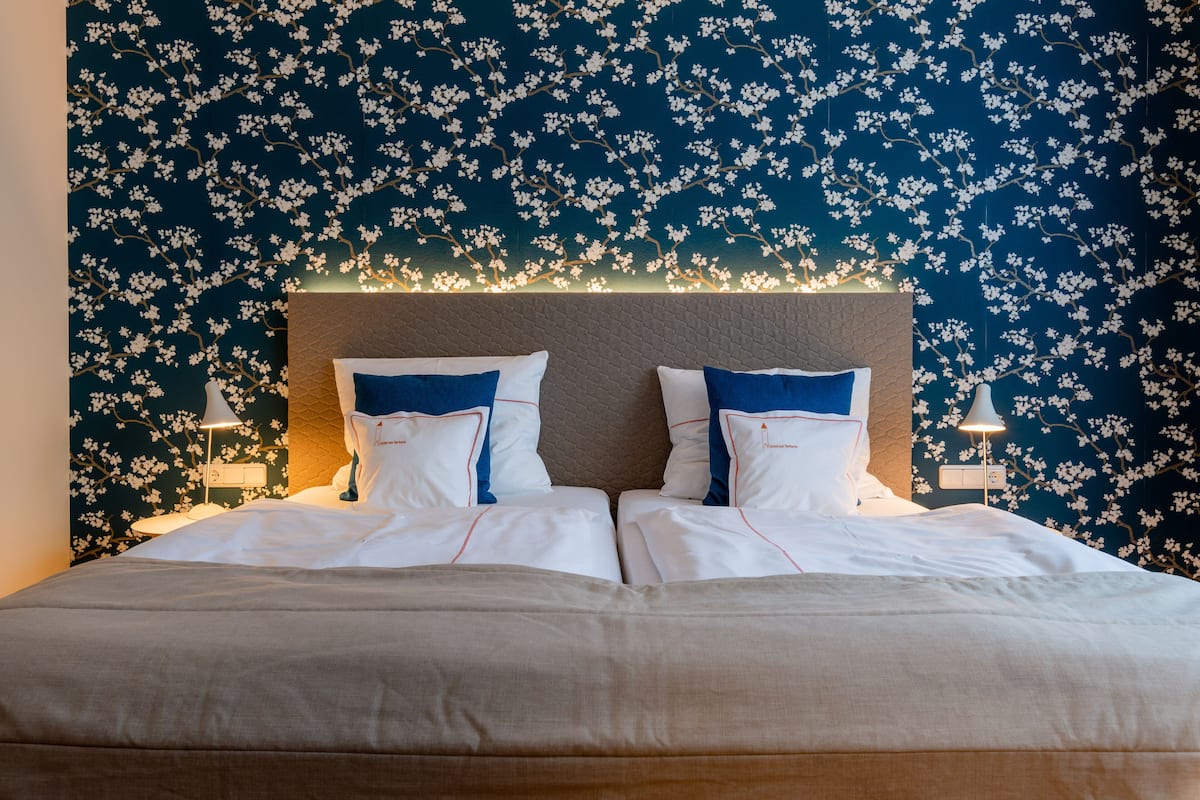 A neatly made bed with white and blue bedding, a headboard with a textured pattern, and a wall with a floral wallpaper design.