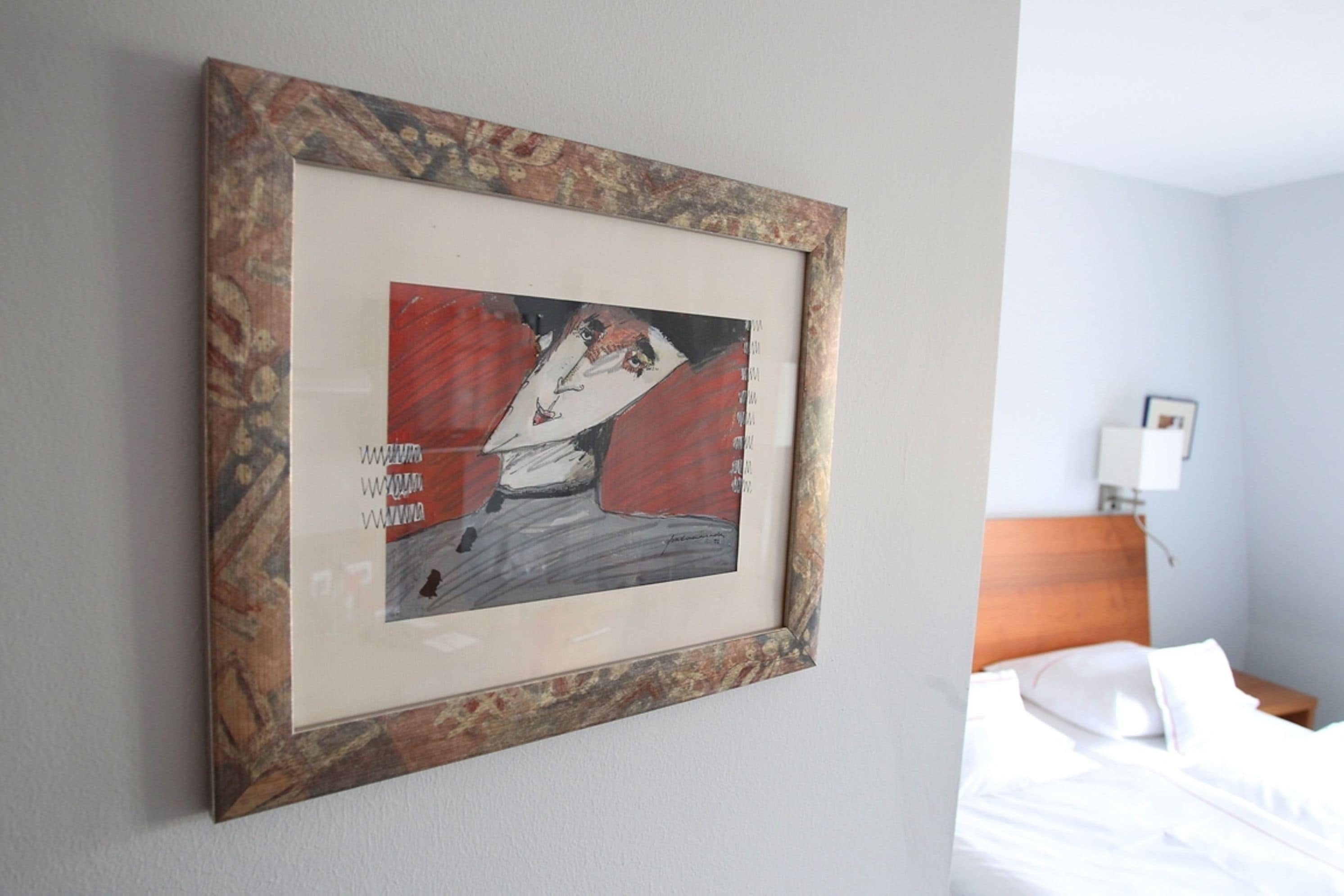 Ein gerahmtes Kunstwerk an einer Hotelzimmerwand, neben einem Bett und einem Nachttisch.