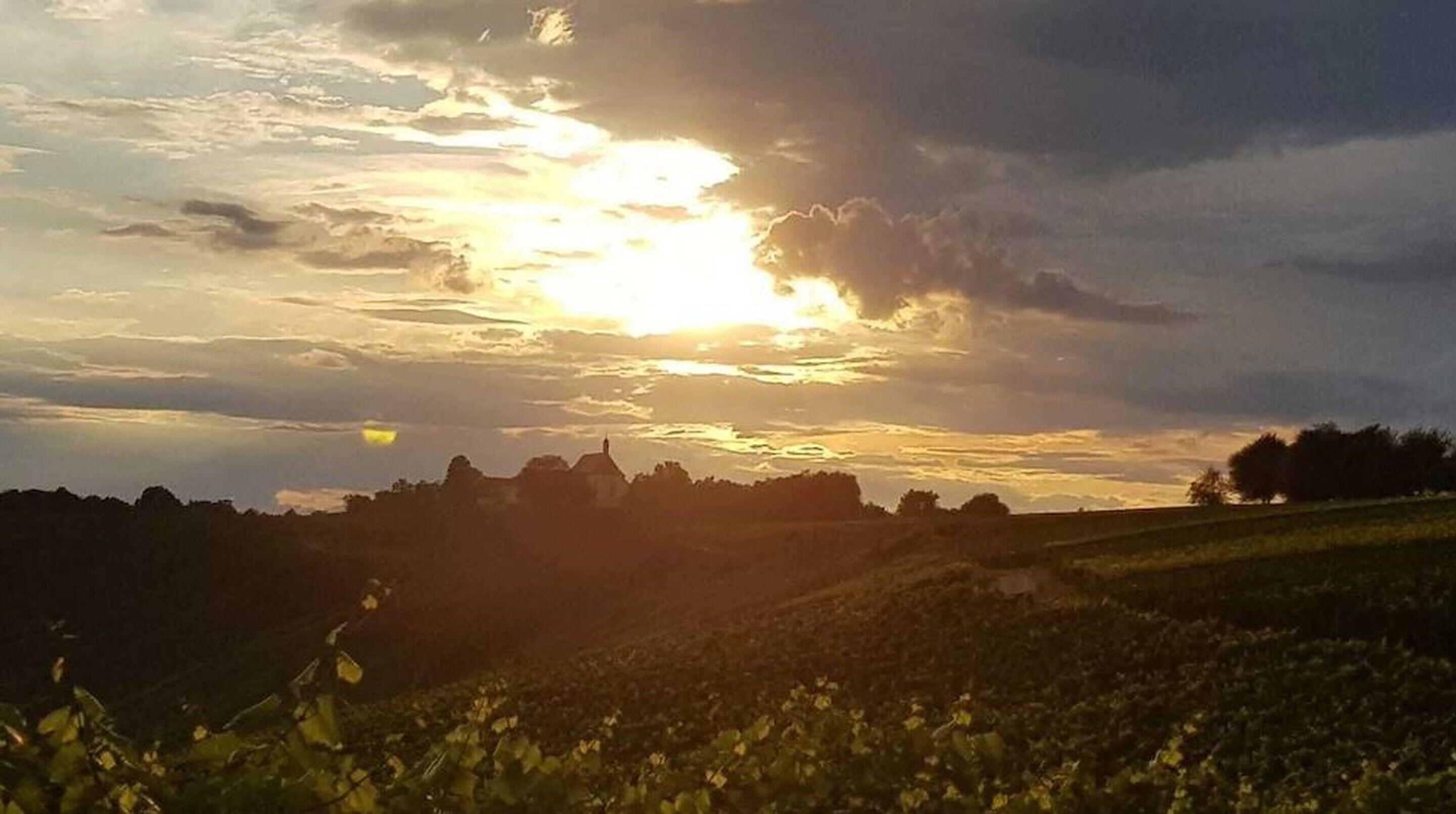 A landscape with a sun setting behind clouds, a hill with vegetation, and a distant building silhouette.