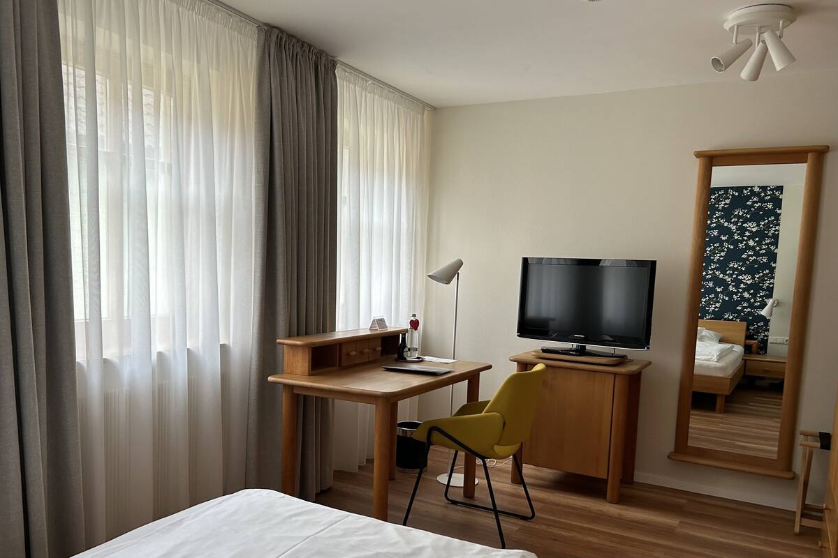 A hotel room with a bed, a desk with a computer, a TV, a chair, a mirror, and a window with curtains.