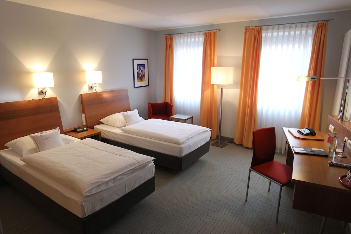 A hotel room with two beds, a desk, a chair, and a lamp.
