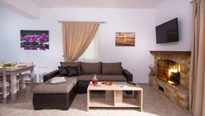 A living room with a sofa, ottoman, coffee table, fireplace, TV, and artwork on the walls. - Elia Villa (Mylopotamos)