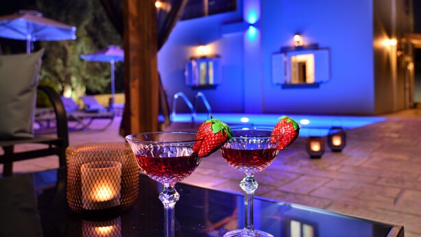 Two cocktails with strawberries on the rim, a lit candle, and a poolside setting with a building in the background. - Elia Villa (Mylopotamos)
