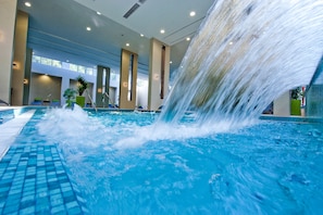 Una piscina interior con una cascada, rodeada de sillones de descanso y plantas en macetas.